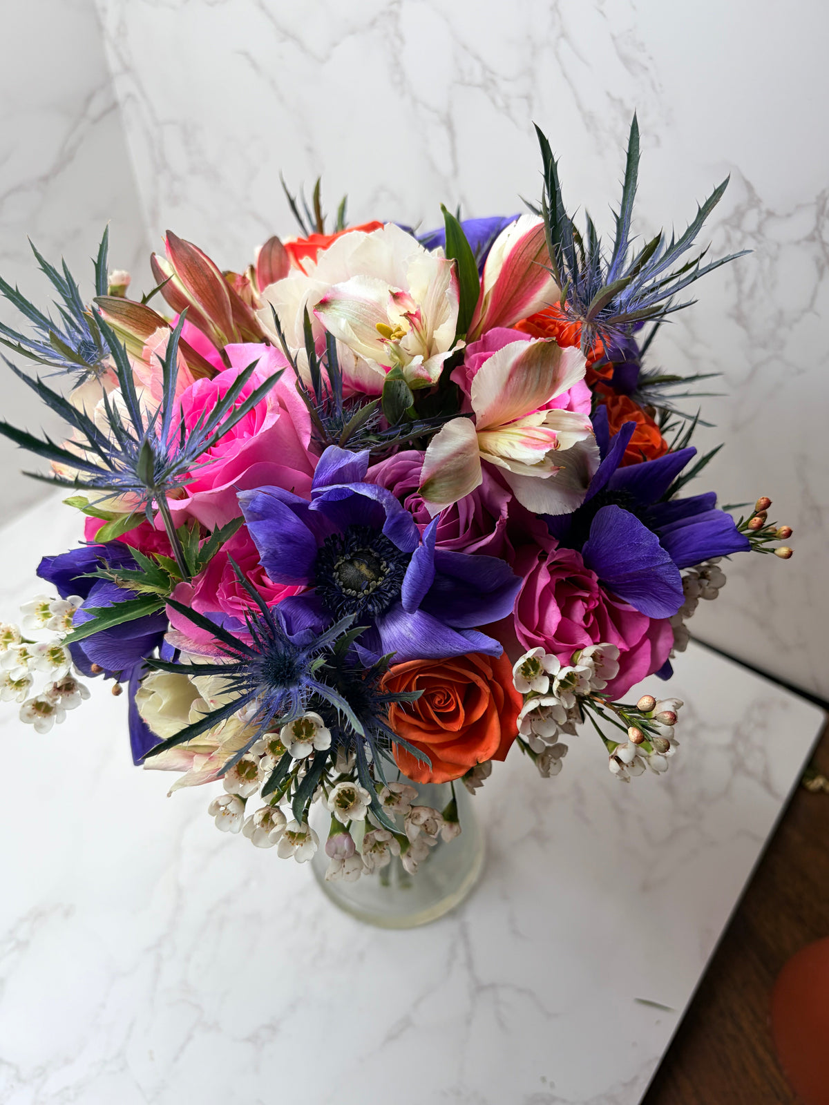 Rose Runners vibrant mixed arrangement with purple anemones, orange roses and blue thistle in a glass vase
